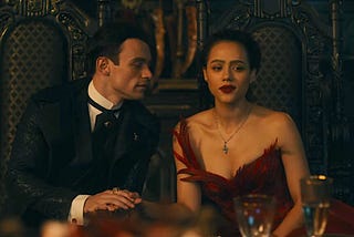 A still frame from the movie The Invitation. We see a handsome aristocrat on the left, holding the hand of a lady in a red dress. He is looking at her, she is looking straight ahead.