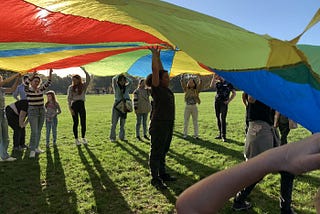 children and student volunteers playing with a rainbow parachute on a sunny day