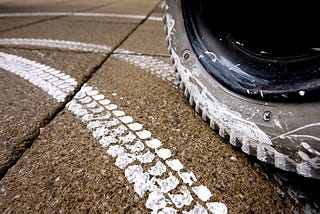 A pavement with a white paint imprint of a wheelchair tire tread. The powered wheel of the wheelchair is visible making the imprint.