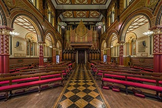 Image of the mid-Victorian chapel with lush red velvet seated wooden pew benches, gilded columns and pillared arches, a large gilded pipe organ at the very back and a pathway patterned tiles.