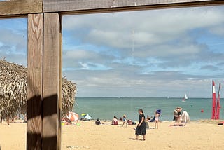 A sunny beach scene with several people relaxing on the sand, a sailboat in the distance, and a thatched hut structure partially visible in the foreground.