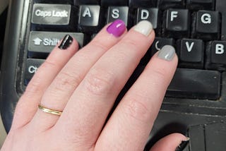 Close up photo of a hand resting on a keyboard. The fingernails are painted in the colours of the asexual flag: black, grey, white, and purple.