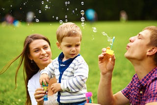 Two young parents play with their toddler with one blowing bubbles and the other holding the child.