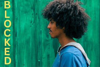 A side on view of a black man with frizzy hair against a green background with the word blocked written in front of him.