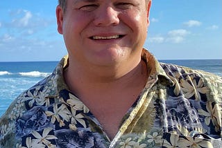 Image of a Keith, a white mane, smiling. He is in Hawaii so you can see the ocean and waves behind him.