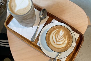 A latte and a cappucino with spoons on the side on a tray on top of a small side table on concrete floor.