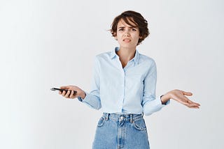 A young woman with short dark brown hair shrugs her shoulders and looks both confused and disgusted while holding a mobile phone.