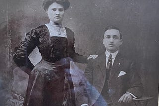 Portrait of the author’s grandmother and grandfather. This photo is from the 1800s, and features a woman in a black long dress, with waist cinched. He’s seated, and wearing a suit.
