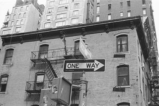 A black-and-white photo of a “one-way sign” in front of a building.
