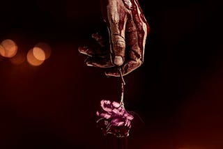 Against a black background, a bloodied hand reaches down, holding what looks like a heart wrapped in wire.