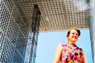 Young woman in a vintage dress, carrying a NIkon camera with a guitar strap under a metallic structure in Europe in the 1980s