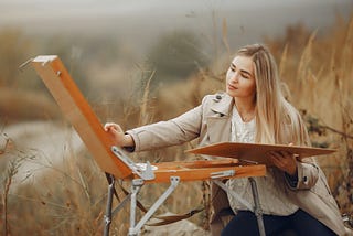 Young woman leaning over a painting box stand, painting a picture of nature outside in a grassy field