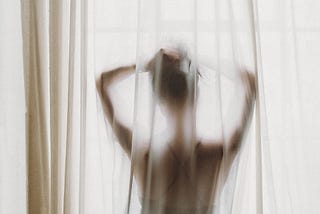 Woman standing behind sheer curtain in front of window.
