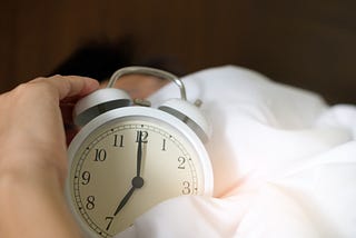 Hand holding analog bell alarm clock in fluffy white bedding with blurry woman sleeping in background.