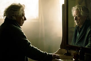 An elderly man stands before a bathroom mirror at dawn while his reflection lingers just slightly behind his movements, creating a subtle but deeply unsettling sense that the two are no longer the same person.