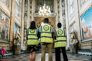 Three people stand, wearing high vis vests with a Night Club logo, with their backs to us in a grand hallway.