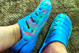 A pair of feet is wearing translucent blue and pink Croc like shoes with multiple cut-outs. The shoes have straps with secure buttons on the sides, and the floor is carpeted.