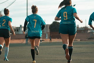 A group of three women wearing teal sports jerseys running on a green field