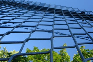 A chain link prison fence with the sky and bushes visable on the other side of it.