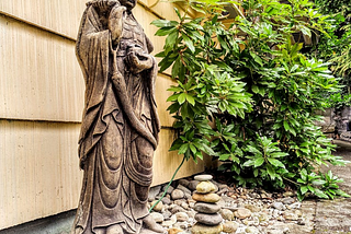 A stand stone statue of Guanyin next to a philodendron. A nine-stone cairn sits at her feet in honor of the author’s mother on her 108th birthday.