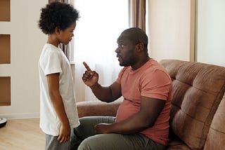 A father sitting on the sofa talking to his son. His finger pointing at his son who is standing in front of him.