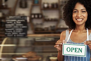 a black woman with an “open” sign on her chest, in front of a coffee shop