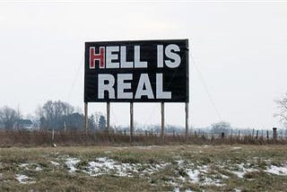 a large black sign sits high about a dead field with patches of snow and dead grass. Barren trees and farmland are in the background. The sky is gray. The big black sign reads in all caps: HELL IS REAL in white block letters with the H in Hell is red.