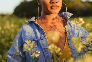 A young woman stands in a field of yellow flowers and stares off into the distance.