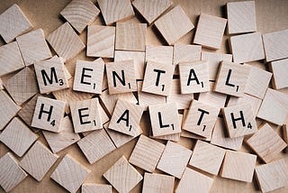 Scrabble tiles scattered on a beige surface spell out ‘MENTAL HEALTH’ with the letters having different point values, conveying the importance and complexity of the topic.