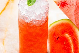 A glass of iced watermelon juice on the counter with cut watermelon next to it.