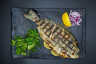 A bbq seared whole fish on a black platter, with chopped, raw red onions, a quarter of a lemon and some parsley sprigs.