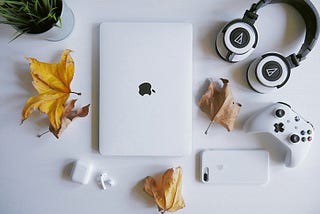 A white MacBook on a white table, surrounded by a potted plant, leaves, AirPods and their case, headphones, an iPhone, and a video game controller.