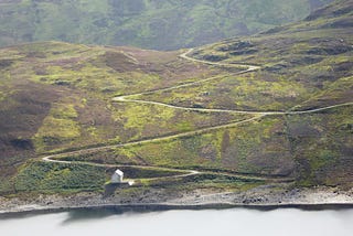 an image of a hill with a zigzag path carved into it, meeting a lake and a small building at the bottom.