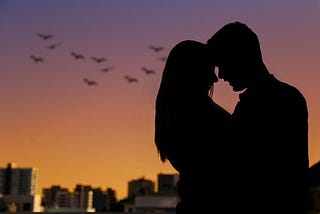 Black sillouette of a heterosexual couple against a dusky sky of orange and purple with birds. She has long hair, their foreheads are touching, they are embracing. Classic fairytale romance… or is it?