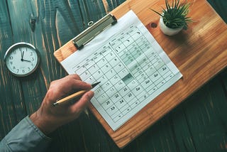 An overhead view of a person’s hand holding a pencil over a printed monthly calendar lying on a wooden desk. Next to the calendar are a silver clipboard clip, a small potted plant, and an analog clock showing approximately 10:09. The desk has a rustic, dark green finish.