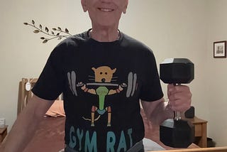 Author smiling while holding a dumbbell in his left hand, wearing a black t-shirt with the words ‘GYM RAT’ and a cartoon design of a rat lifting weights. He is standing in a bedroom with a wooden bed frame and decorative wall art in the background.