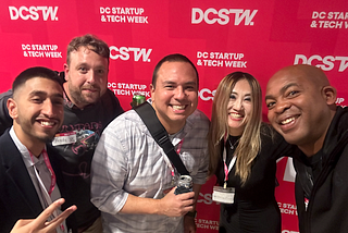 A group of five people posing and smiling at DC Startup & Tech Week in front of a red backdrop with the event logo. From left to right, a man in a suit makes a peace sign, the second person (wearing a Brady UX name tag) stands in a graphic tee, the third person is holding a drink and smiling, the fourth person is a woman with long hair, and the fifth person is smiling broadly. They appear to be enjoying the networking atmosphere of the event.