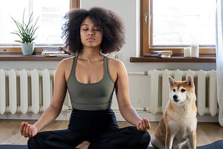 A young Black woman sitting on a yoga mat in lotus pose. A spitz dog sits at her side.