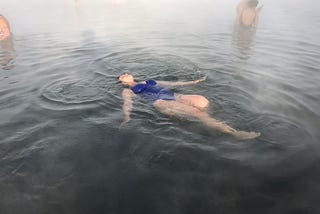 Woman on her back in a blue bathing suit with leg outstretched and arms at her sides in the middle of a hot spring.