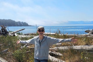 Author, Gill McCulloch on the beach in Sooke, BC, Canada against a background of blue sky, ocean and trees. In the foreground are driftwood logs and shrubs.