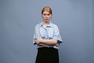 A young woman with red hair wearing a blue, button-up shirt and black skirt with a determined expression.