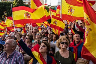 Photo of people waving Spanish flags during a protest demanding general elections and against Pedro Sanchez’s Government after motion of censure, in Madrid, Spain. The crowd is looking towards the left, with a few people looking towards the right in the bottom corner of the photo.