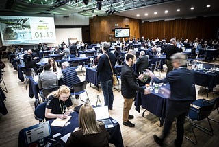 A large crowded room filled with tables for various meetings, and a projector reflects a clock ticking to direct the attendees when to move to their next meeting.