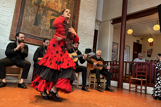 A female flamenco dancer in a frilly red and black dress dances in front of four male singers and guitarists.