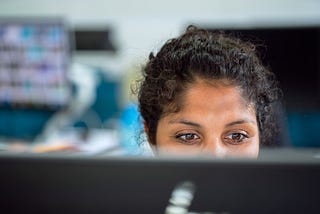 A woman with curly black hair that is pulled back, sits in a computer lab with her eyes peeking over a computer screen.