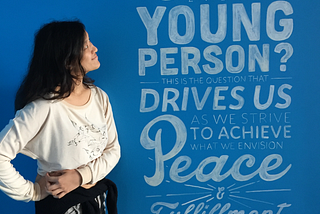 woman looking at a wall with text written “can you imagine what will happen to the world when we engage and develop every young person? that is the question that drives us to achieve peace and fulfillment of humankind’s potential”