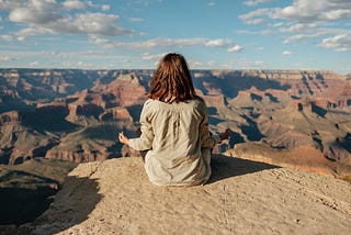 Long-haired person sitting facing a natural view with their back to the camera, in a meditative position.