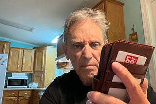 An older man standing in a kitchen, looking intently and somewhat frustrated or even angry at his smartphone, which is in a leather wallet-style case. The background shows wooden cabinets, a microwave, and a bright overhead light.