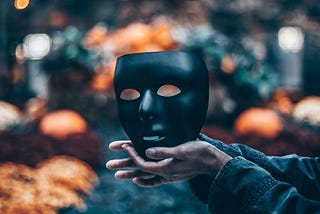 A completely black face mask being held, cupped in two hands before a blurry colorful background.