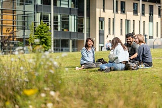 A group of four students sit cross legged on grass outside the Michael Marks building. They are all dressed in light summer clothing. and are smiling and in conversation. There is greenery in the foreground out of focus. The building is modern architectural style.